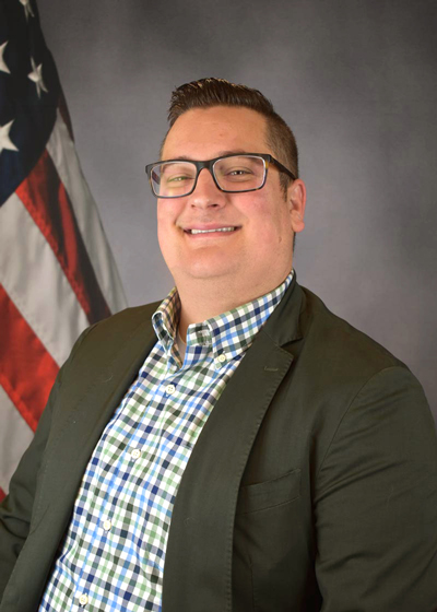 Picture of Sean Cook in a green and blue plaid button up shirt and green blazer sitting in front of a grey backdrop with an American flag.